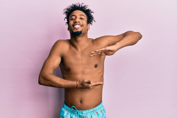 Junger afrikanisch-amerikanischer Mann mit Bart im Badeanzug gestikuliert mit Händen, die große und große Zeichen zeigen, Maßsymbol. Lächelnd in die Kamera blickend. Messkonzept.  - Foto, Bild