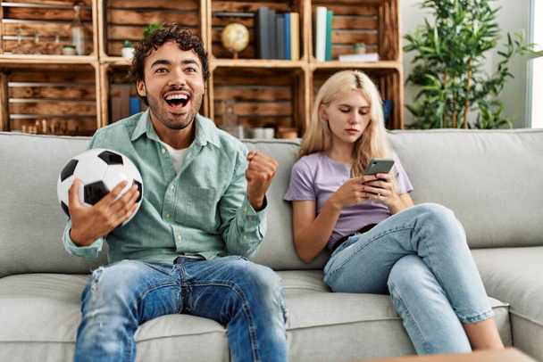 Mężczyzna uśmiechnięty szczęśliwy oglądając mecz piłki nożnej i dziewczyna nudne za pomocą smartfona w domu. - Zdjęcie, obraz