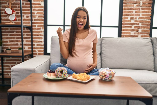 Mujer embarazada joven comiendo dulces en casa celebrando la victoria con sonrisa feliz y expresión ganadora con las manos levantadas  - Foto, imagen