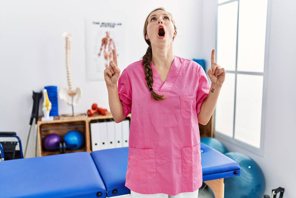 Ağrı giderme kliniğinde çalışan genç sarışın kadın şaşkın ve şaşırmış bir şekilde yukarı bakıyor ve parmaklarıyla işaret ediyor ve kollarını kaldırıyor..  - Fotoğraf, Görsel