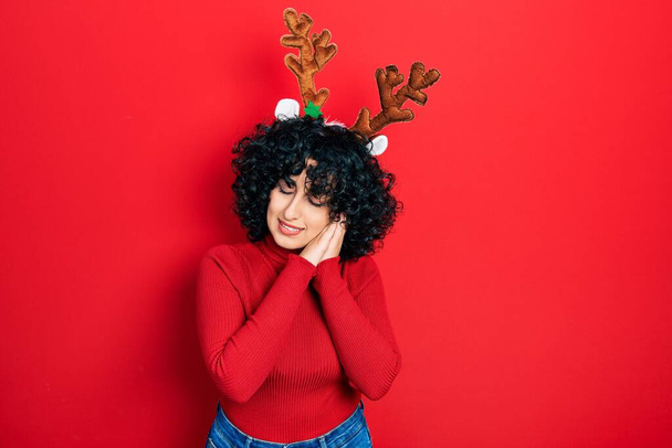 Joven mujer de Oriente Medio vistiendo lindos cuernos de renos navideños durmiendo cansada soñando y posando con las manos juntas mientras sonríe con los ojos cerrados.  - Foto, Imagen
