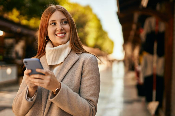 Νεαρή κοκκινομάλλα κορίτσι χαμογελώντας ευτυχισμένη χρησιμοποιώντας smartphone στην πόλη. - Φωτογραφία, εικόνα