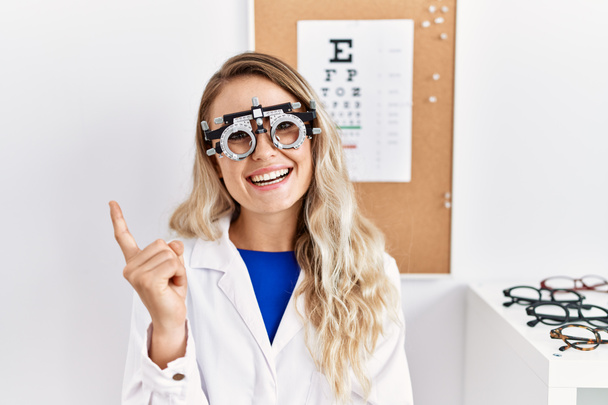 Молодая красивая оптик женщина в очках оптометрии в клинике улыбается счастливо указывая рукой и пальцем в сторону  - Фото, изображение