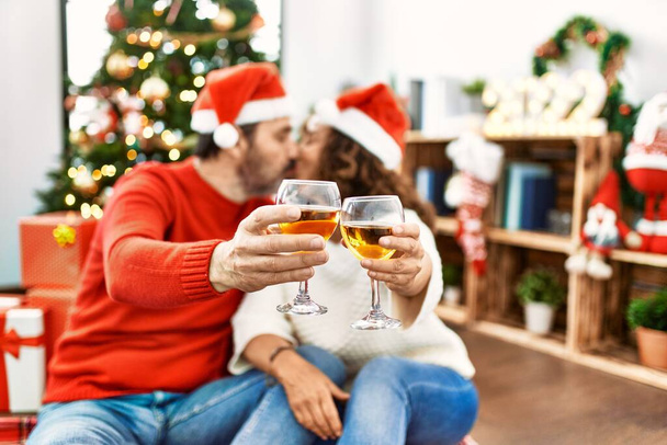 Casal hispânico de meia-idade com chapéu de Natal. Sentado no chão brindando com champanhe em casa. - Foto, Imagem