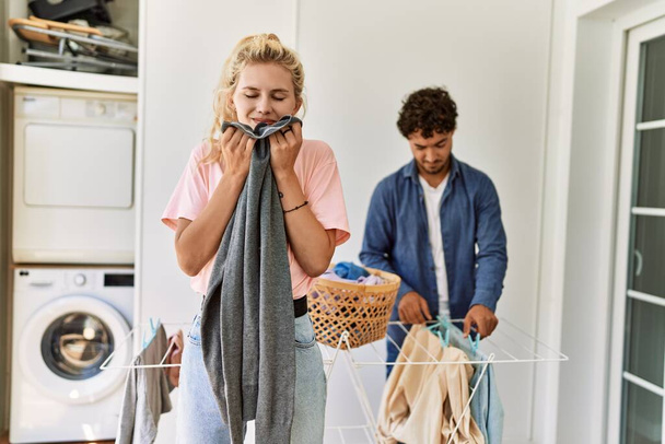 Ένα νεαρό ζευγάρι χαμογελά χαρούμενο κάνοντας μπουγάδα. Γυναίκα μυρίζει καθαρά ρούχα στο σπίτι. - Φωτογραφία, εικόνα