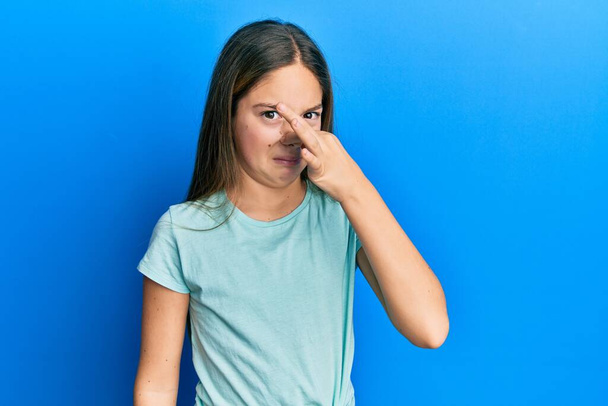 Beautiful brunette little girl wearing casual white t shirt smelling something stinky and disgusting, intolerable smell, holding breath with fingers on nose. bad smell  - Photo, Image