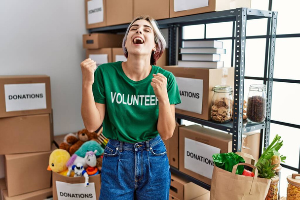 Young beautiful woman wearing volunteer t shirt at donations stand very happy and excited doing winner gesture with arms raised, smiling and screaming for success. celebration concept.  - Foto, immagini