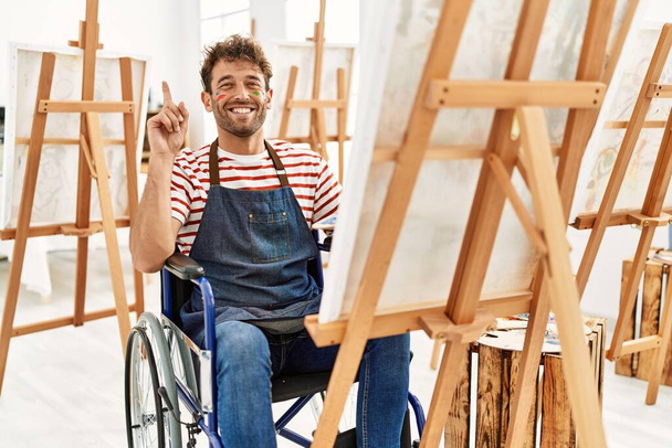 Young handsome man with beard at art studio sitting on wheelchair surprised with an idea or question pointing finger with happy face, number one  - 写真・画像