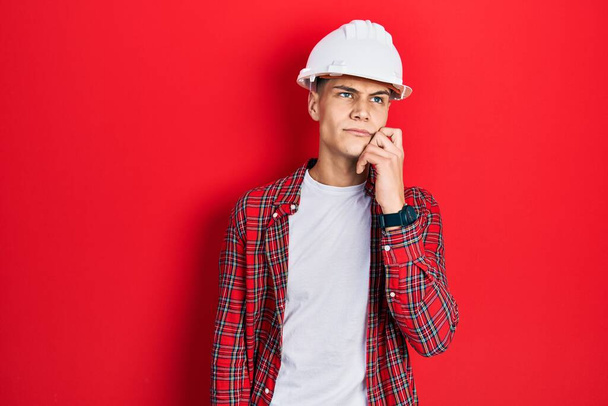 Young hispanic man wearing architect hardhat serious face thinking about question with hand on chin, thoughtful about confusing idea  - Photo, image