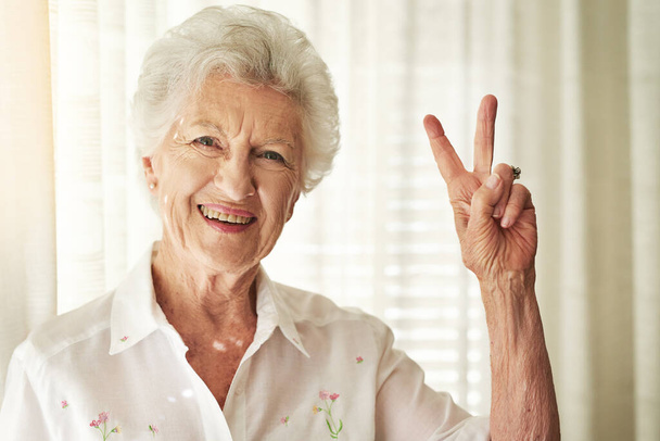 V jak witalność. Portret szczęśliwej starszej kobiety okazującej gest pokoju w domu. - Zdjęcie, obraz