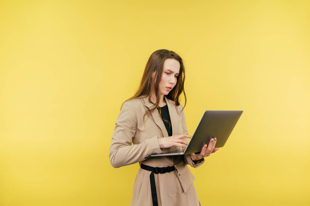 Kaunis naispuolinen toimistotyöntekijä takissa seisoo keltaisella taustalla ja käyttää kannettavaa tietokonetta, jolla on vakavat kasvot. - Valokuva, kuva