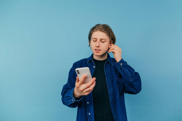 Θετικός νεαρός άνδρας σε ασύρματα ακουστικά απολαμβάνει smartphone ακούγοντας μουσική με σοβαρό πρόσωπο σε μπλε φόντο. - Φωτογραφία, εικόνα