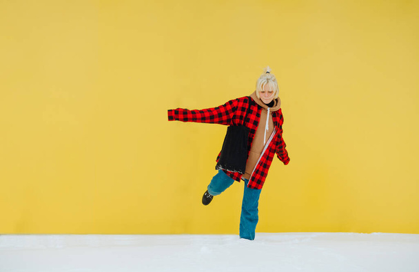 Lustiges Teenie-Mädchen in lässiger Kleidung, das im Winter im Freien Spaß hat und auf einem Bein im Schnee vor einer gelben Wand steht. - Foto, Bild