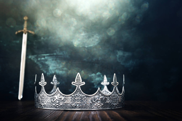 low key image of beautiful queen or king crown over antique next to sword. fantasy medieval period. Selective focus. Glitter sparkle lights - Photo, Image