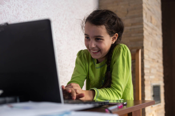 Yüzünde kulaklıkla gülümseyen küçük beyaz kız bilgisayarını kullanarak uzaktan öğretmeniyle video görüşmesi yapıyor, öğretmeniyle mutlu küçük çocuk dalgası selamlaması yapıyor, bilgisayarda çalışıyor, evde eğitim kavramı geliştiriyor. - Fotoğraf, Görsel