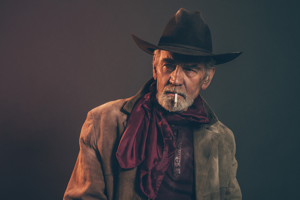 Velho cowboy ocidental áspero com barba cinza e chapéu marrom fumando um
 - Foto, Imagem