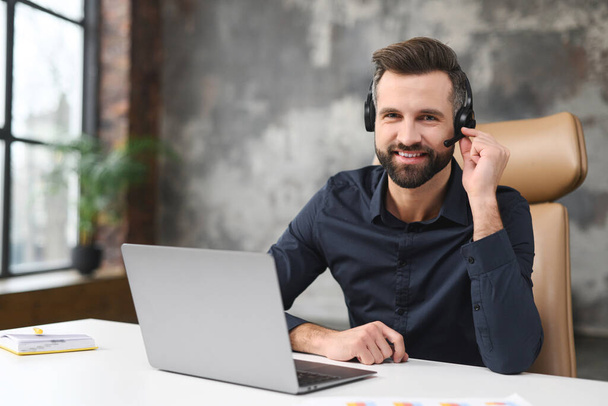 Friendly young guy in smart casual wear is using headset and laptop for online communication, supporting, selling. Guy sitting at the office desk and smiling - Foto, Bild
