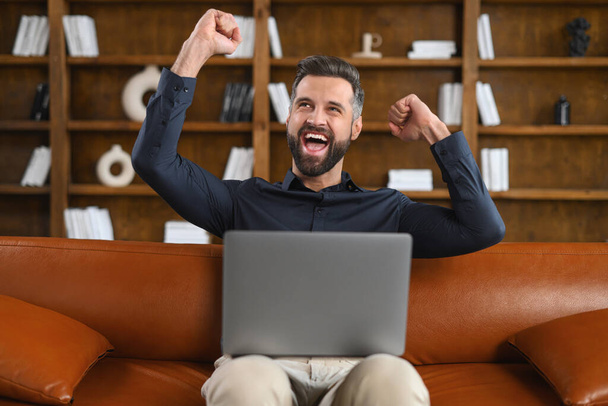 Excited man sitting on the brown sofa looking away in front of the laptop and screaming, surprised emotional lucky man with wide opened mouth - Zdjęcie, obraz