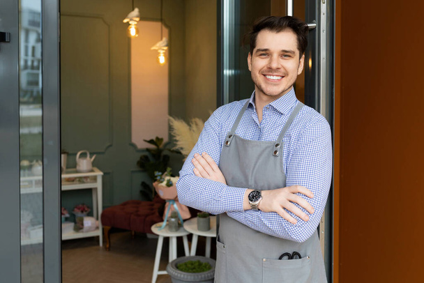 Владелец кафе с широкой улыбкой рядом с входом в свой ресторан в фартуке приветствует клиентов - Фото, изображение