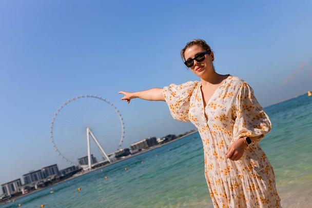 Счастливые женщины-туристки прогуливаются босиком по песчаному пляжу в районе аэропорта Дубая и любоваются панорамным видом на колесо обозрения Айн Феррис - Фото, изображение