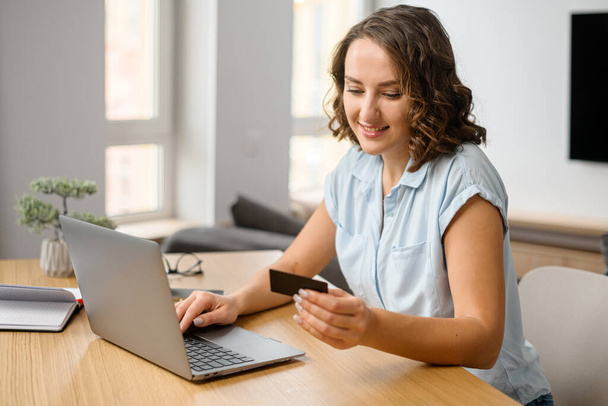 Smiling caucasian teenage girl sitting at the laptop, entering credit card number to make an online transaction. Woman ordering food - Foto, afbeelding