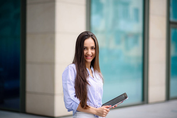 Young cheerful business lady in earphones looking at digital tablet with smile while standing on sunny street outdoors, listening webinar or participating in online business meeting with team - Φωτογραφία, εικόνα