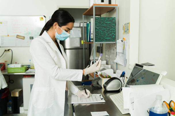 Vrouwelijke biochemicus die de patiëntinformatie schrijft terwijl hij een reageerbuis vasthoudt met een bloedmonster in het medisch lab - Foto, afbeelding