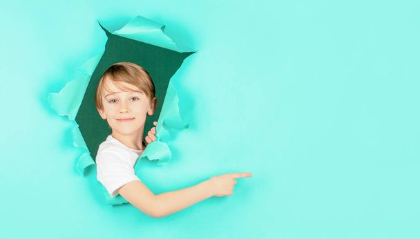 Kid looks through hole. Blonde boy looks through ripped paper. Boy in red paper hole pointing with finger aside. Smile boy looking through hole in paper wall. Surprised child making hole in paper - Fotó, kép