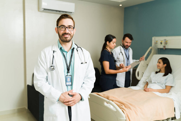 Przystojny lekarz uśmiechający się i patrzący w kamerę, podczas gdy personel medyczny sprawdza chorego pacjenta w szpitalnym łóżku - Zdjęcie, obraz