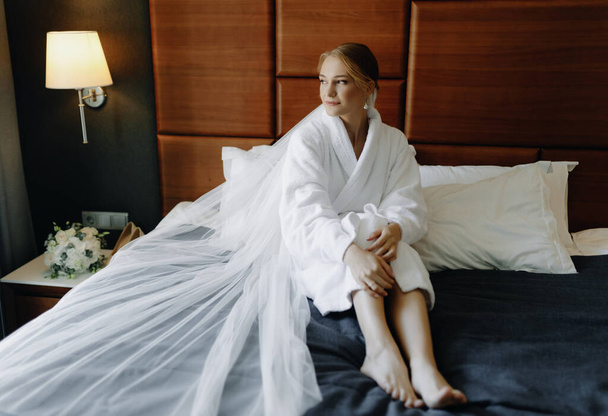  La sposa in accappatoio bianco con velo si siede sul letto e distoglie lo sguardo. Foto di alta qualità - Foto, immagini