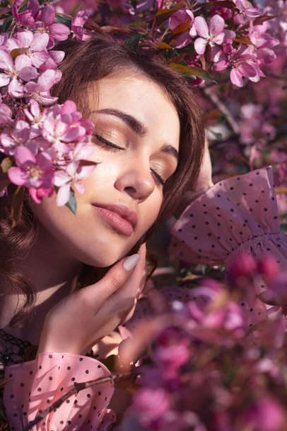 Retrato de uma jovem menina bonita com olhos fechados com cabelos castanhos longos em um vestido rosa em um fundo de flores de cereja. - Foto, Imagem