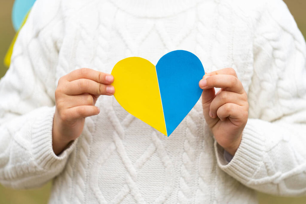 Ένα μικρό κορίτσι κρατά στα χέρια της κοντά στο στήθος της μια καρδιά στα πατριωτικά χρώματα της ουκρανικής σημαίας, κίτρινο και μπλε. Σταματήστε τον πόλεμο στην Ουκρανία. Βοήθησε να σωθεί το Ουκρανικό έθνος. Υψηλής ποιότητας φωτογραφία - Φωτογραφία, εικόνα