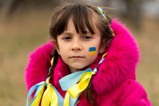 A little Ukrainian refugee girl with a Ukrainian flag painted on her cheek is sad about her lost home. Stop the war in Ukraine. Help save the Ukrainian nation. High quality photo - Valokuva, kuva