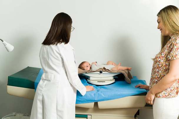 Tylny widok pediatry ważącego zdrowe dziecko. Biała mama przynosząca dziecko na badania lekarskie w szpitalu - Zdjęcie, obraz