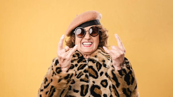 Stylish senior woman showing rock n roll gesture while posing on isolated background. - Фото, изображение