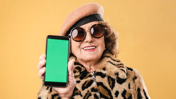 Κομψή ηλικιωμένη γυναίκα που δείχνει ένα κινητό τηλέφωνο με πράσινο ενώ στέκεται πάνω από ένα απομονωμένο φόντο. - Φωτογραφία, εικόνα