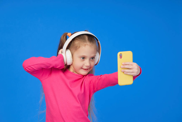 A cute girl in a bright pink sweater taking a selfie on a mobile phone camera and listens to music in wireless headphones. The concept of children's applications on gadgets - Zdjęcie, obraz