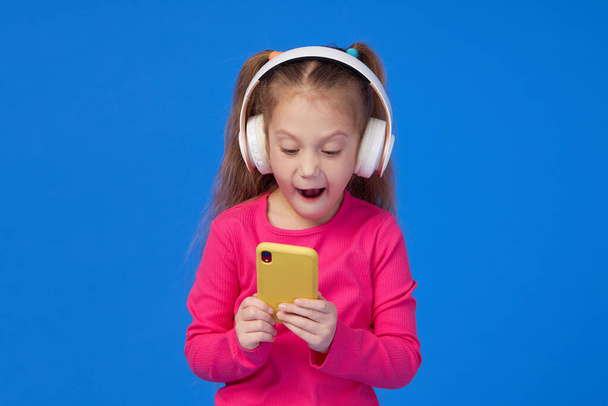 A cute girl in a bright pink sweater holds a mobile phone in her hands and listens to music in wireless headphones. The concept of children's applications on gadgets - 写真・画像