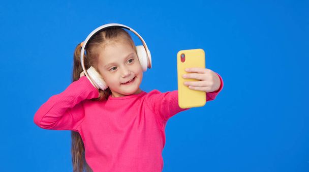 A cute girl in a bright pink sweater taking a selfie on a mobile phone camera and listens to music in wireless headphones. The concept of children's applications on gadgets - Foto, Bild