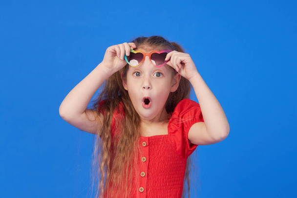Portrait of surprised cute little toddler girl in the heart shape sunglasses. Child with open mouth having fun isolated over blue background. Looking at camera. Wow funny face - Photo, image