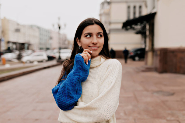 Romantic European woman with lovely smile and dark hair is wearing spring bright knitted sweater is looking up and holding hand on the lips. Pretty woman is walking on the street in warm spring day - Photo, Image