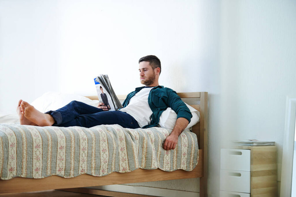Потрапити в трішки новин. Знімок красивого молодого чоловіка, що лежить на ліжку, читаючи журнал
. - Фото, зображення