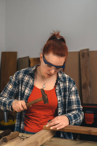 νεαρή κοκκινομάλλα, ξυλουργός σφυρηλατεί ένα καρφί σε μια ξύλινη σανίδα στο μικρό ξυλουργείο της. - Φωτογραφία, εικόνα