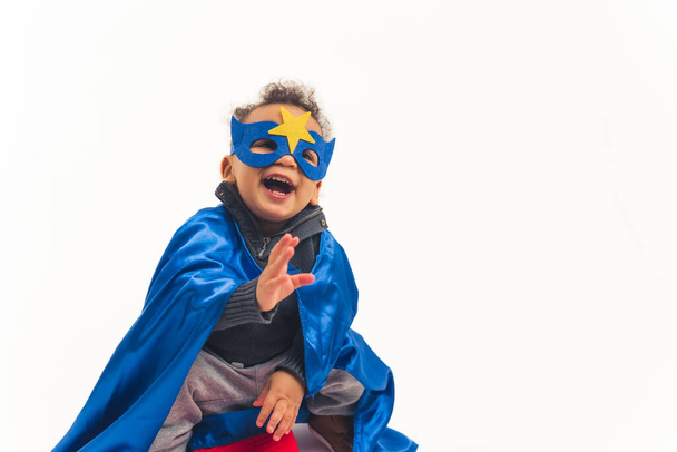 Studio shot of a little biracial boy toddler in a hero costume with a dark blue mask with a star and a cape laughing out loud over white background. - Photo, Image
