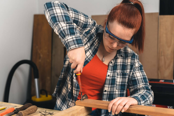 Jovem mulher de cabelos vermelhos carpinteiro aparafusar um parafuso em uma prancha de madeira. - Foto, Imagem