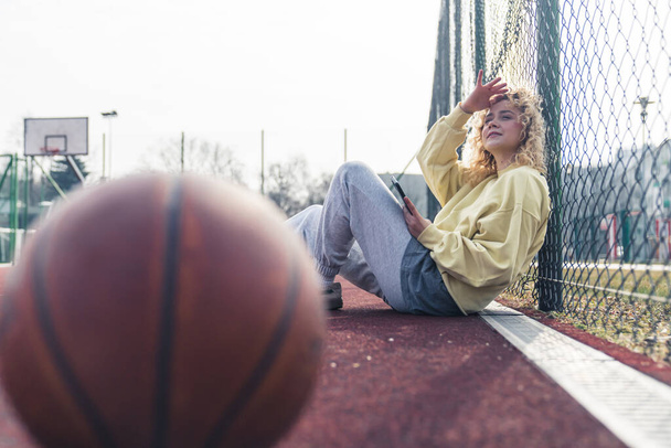 Όμορφη νεαρή λευκή γυναίκα κάθεται στο γήπεδο του μπάσκετ, κρατώντας το τηλέφωνο και κοιτάζοντας προς τα πάνω, η μπάλα του μπάσκετ είναι στο μπροστινό αντίγραφο χώρο  - Φωτογραφία, εικόνα
