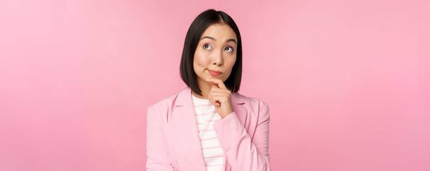 Image of asian businesswoman standing in thinking pose, brainstoming, wearing suit. Korean saleswoman, entrepreneur posing against pink background - 写真・画像