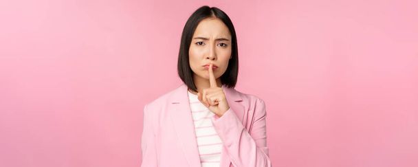 Тихо, табу концепция. Портрет азиатской предпринимательницы, показывающей молчаливый жест, шшшш знак, прижимающей палец к губам, стоящей на розовом фоне в костюме - Фото, изображение