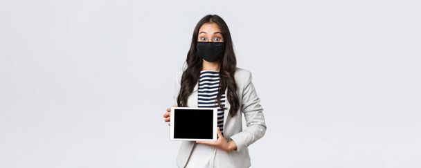 Wirtschaft, Finanzen und Beschäftigung, covid-19 Prävention Virus und soziale Distanzierung Konzept. Aufgeregte asiatische Geschäftsfrau mit Gesichtsmaske und Brille öffnet erstaunt die Augen und zeigt ein digitales Tablet-Display - Foto, Bild