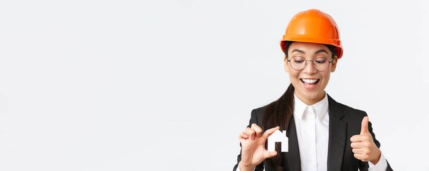 Close-up of happy pleased asian female architect, construction engineer in helmet and suit looking at home miniature and showing thumbs-up, guarantee quality of repairment works, white background - Photo, Image